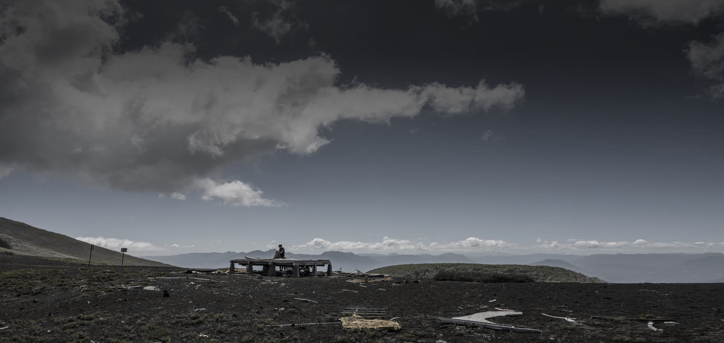 A lone figure sits on a collapsed wooden platform on a dark volcanic plain beneath a heavy grey sky, mountains on the horizon.